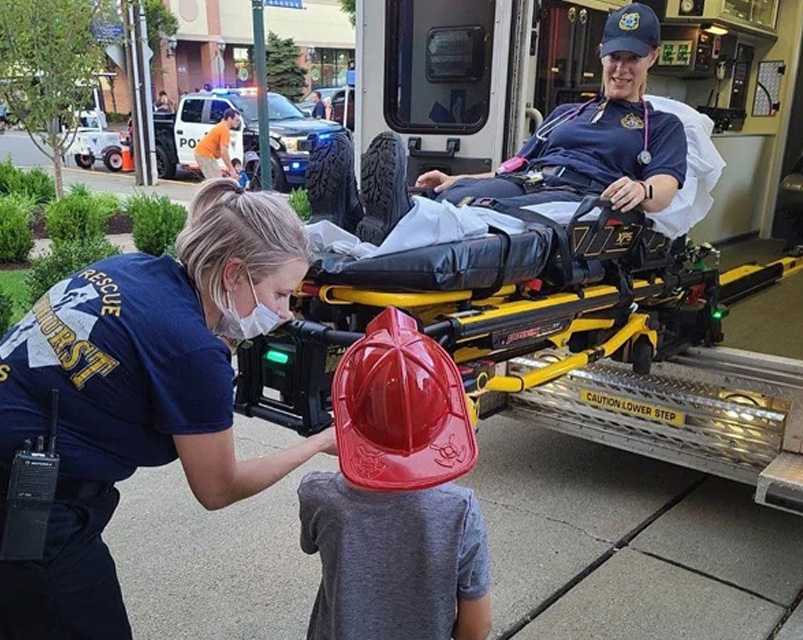 ambulance-stats LPES EMT greets a child wearing a red firefighter helmet beside an ambulance stretcher.